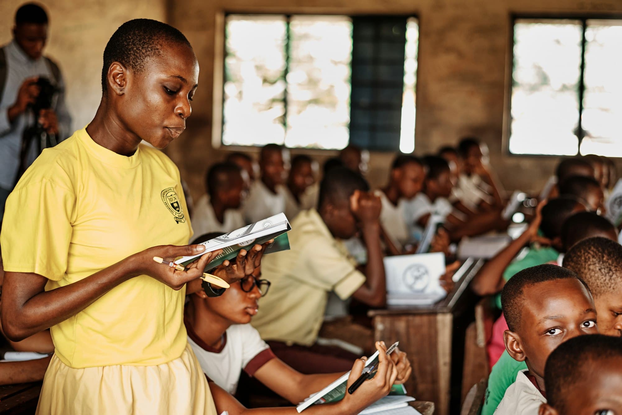 A student in a yellow school shirt reads aloud from a book while classmates follow along
